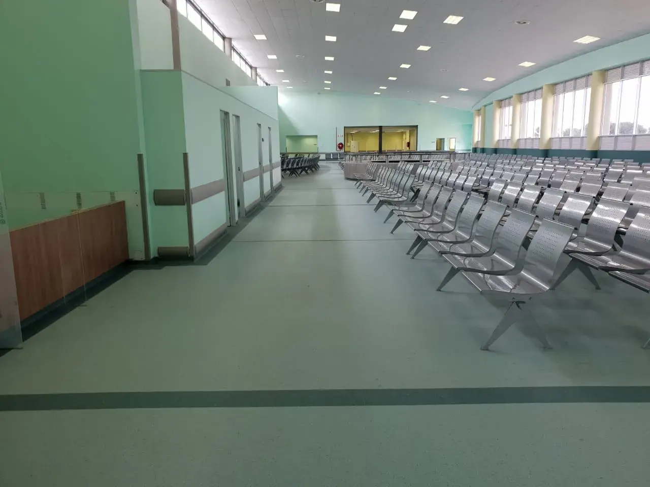 A large clinic waiting hall with rows of steel seating on a green vinyl floor with a dark inset line.