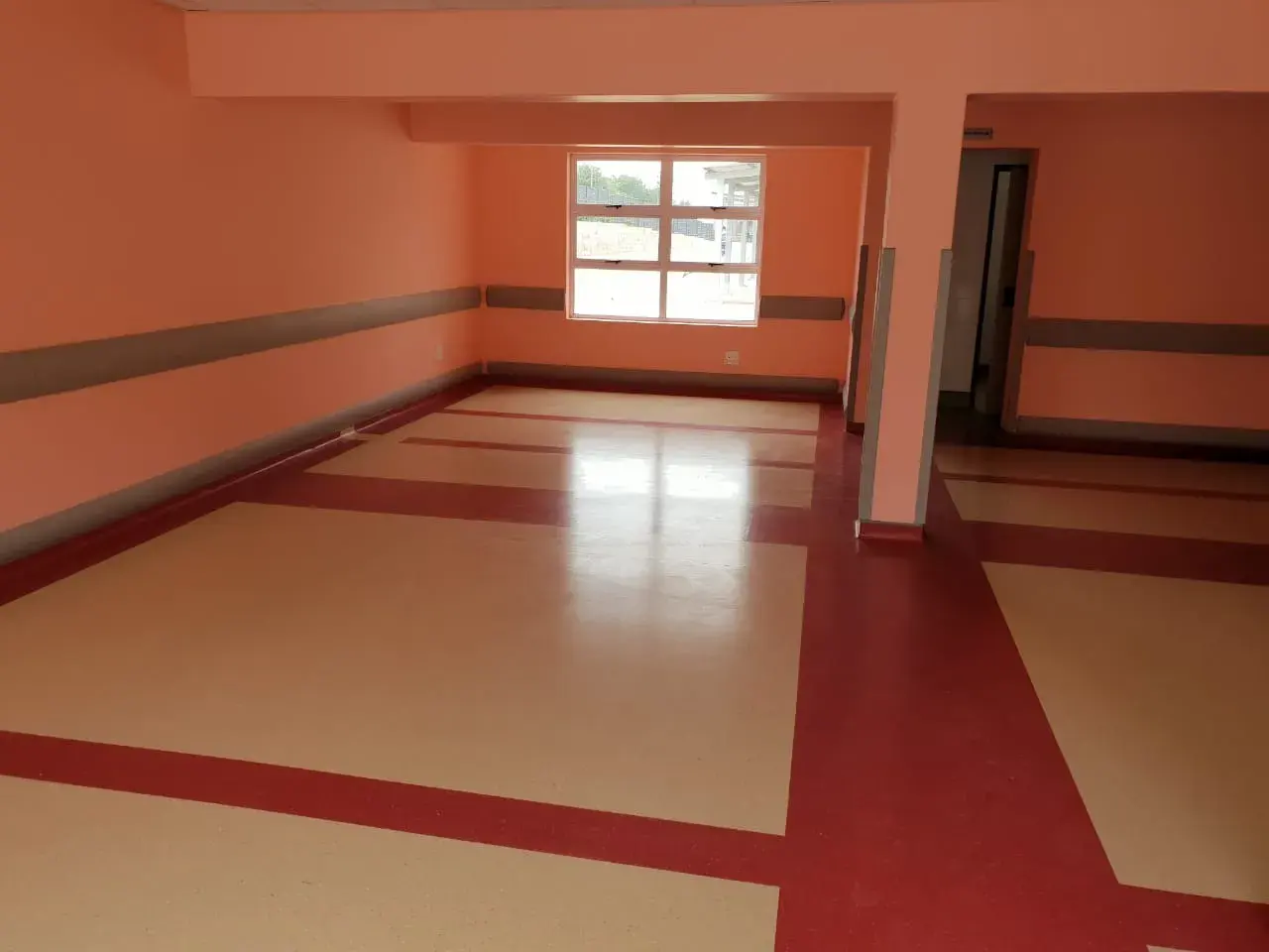 An empty clinic ward floored in cream vinyl with deep red borders inlaid around the bed bays.
