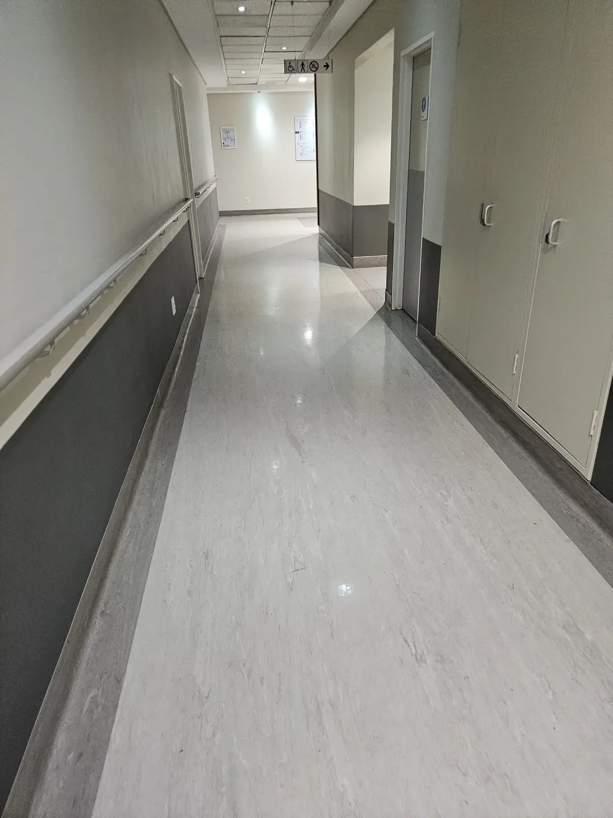 A long hospital corridor in light grey sheet vinyl with a darker border, a handrail and dark grey wall protection.