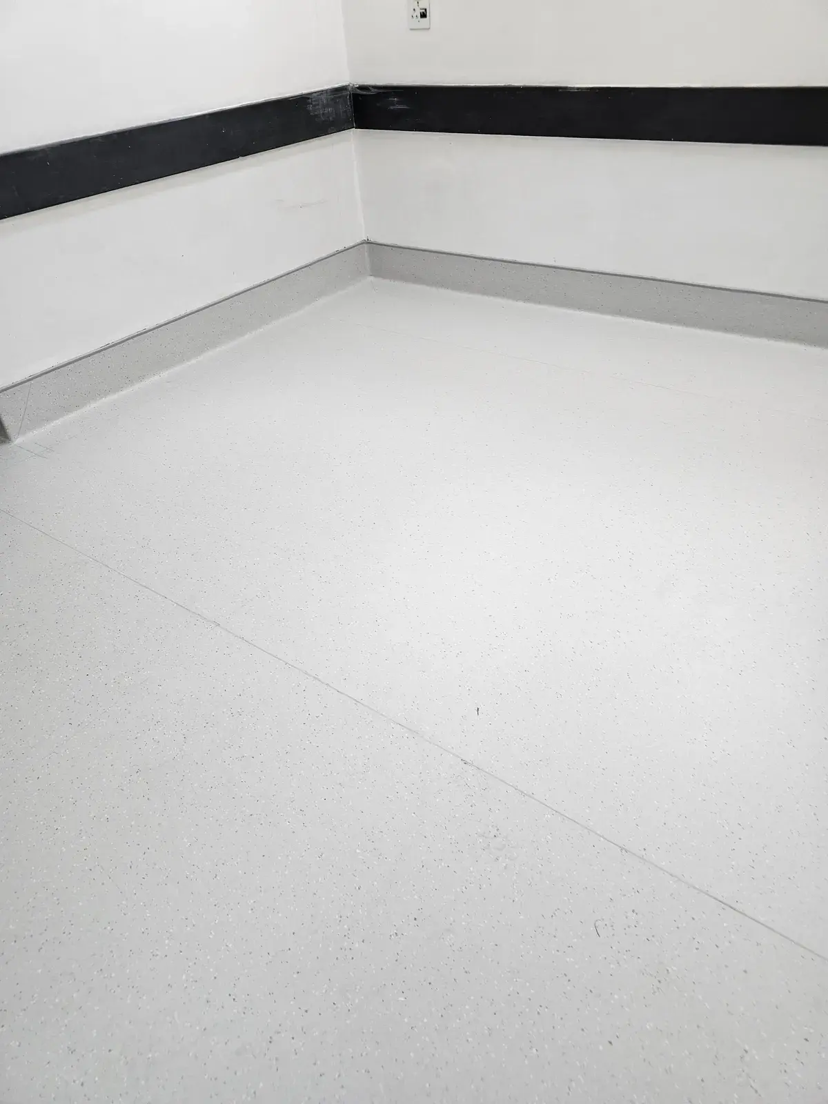The corner of a room in white speckled vinyl, with a grey coved skirting and a black wall guard.