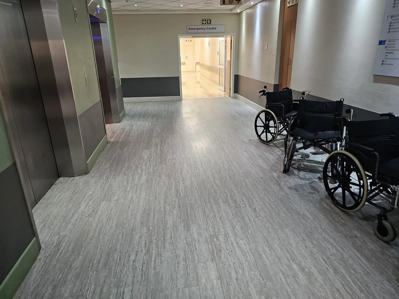 A hospital lift lobby with a grey vinyl floor, wheelchairs parked along the wall and the emergency centre entrance ahead.